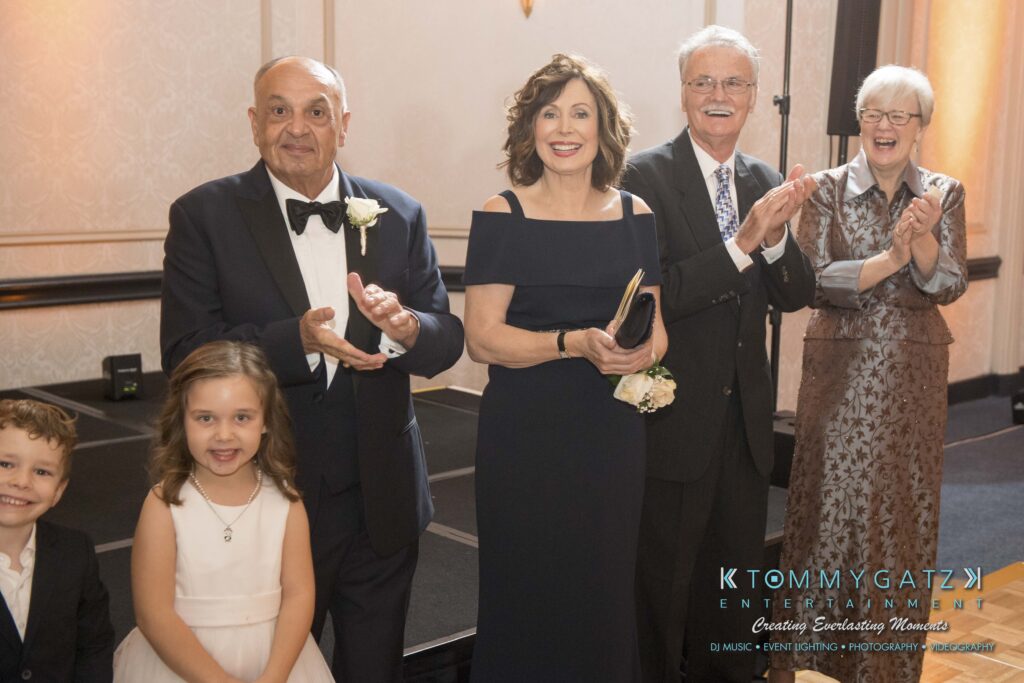 A group of five smiling adults, dressed formally, standing in a line with two children in front. They're in an event space with subtle lighting.