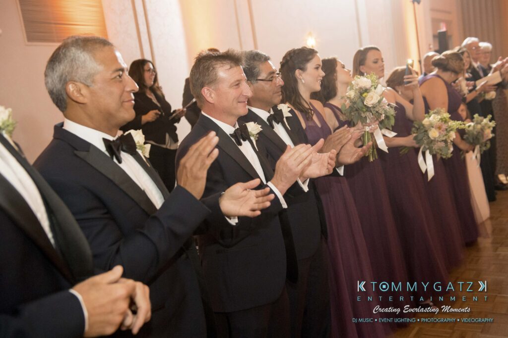 A group of people in formal attire, including men in tuxedos and women in matching purple dresses, stand in a row clapping at an indoor event.