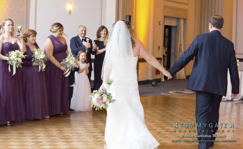 Bride and groom walk hand in hand on dance floor, bridesmaids in purple dresses watching.