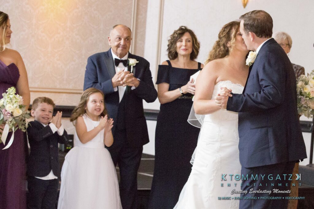 A bride and groom kiss at a wedding ceremony. A group of people, including children and an elderly man, watch and clap. A floral arrangement is in the foreground.
