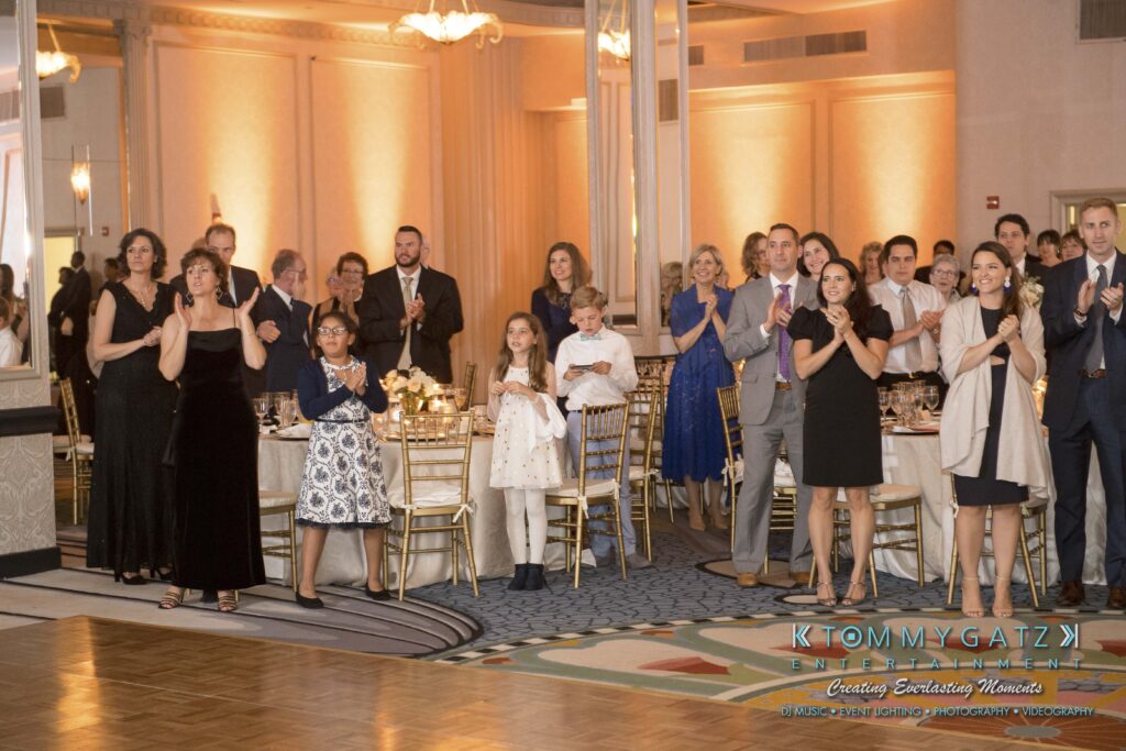 Guests at a formal event standing and clapping, with tables set elegantly and decorations visible.