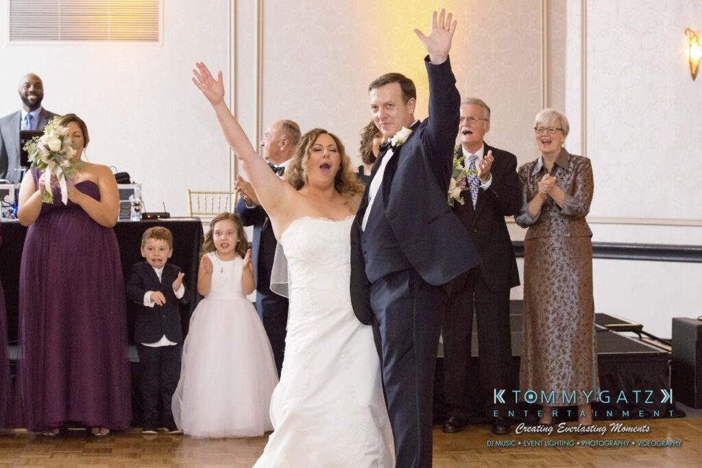 Bride and groom celebrate with raised hands at a wedding reception, surrounded by smiling guests and children.