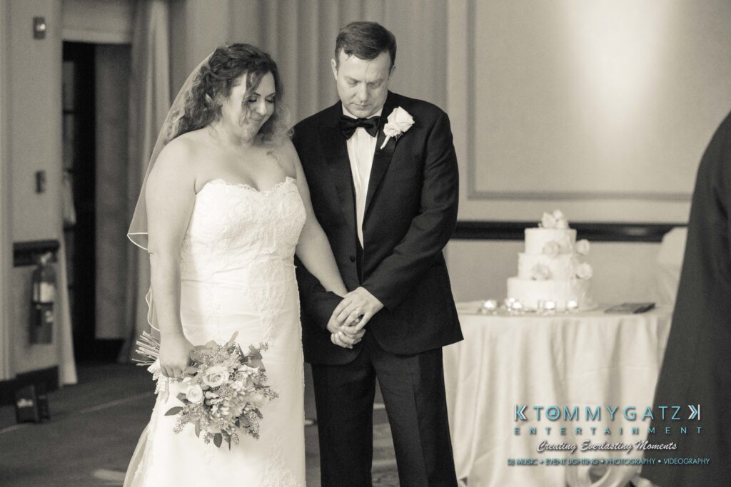 A bride and groom hold hands in a wedding ceremony. The bride holds a bouquet, and a wedding cake is visible in the background.