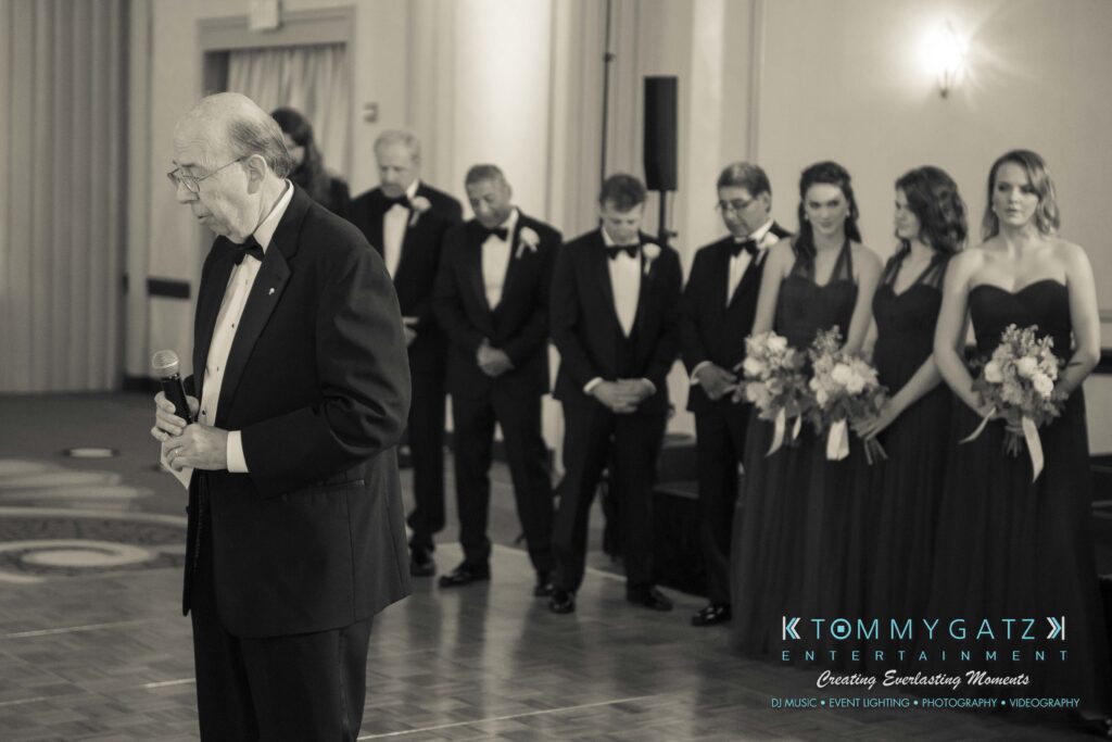 An elderly man in a suit speaks into a microphone. A group of people in formal wear stands in the background, some holding bouquets.