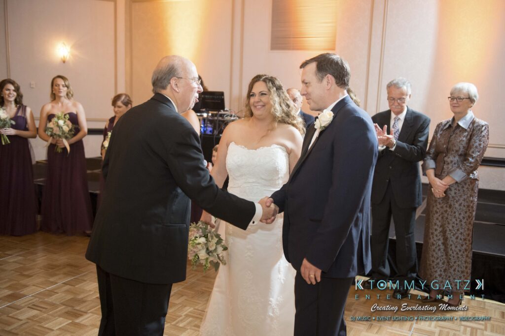 A bride in a white dress smiles as a man in a black suit shakes hands with a groom in a blue suit during a wedding ceremony, with bridesmaids and guests in the background.
