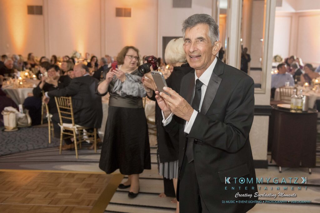 Man in a suit smiling and taking a photo with a smartphone at an indoor event. People seated and standing in the background.