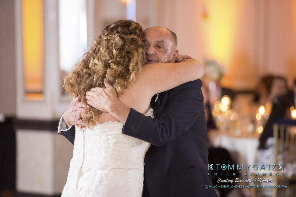 A bride and an older man share a dance at a formal event. The room is elegantly decorated with warm lighting and round tables.