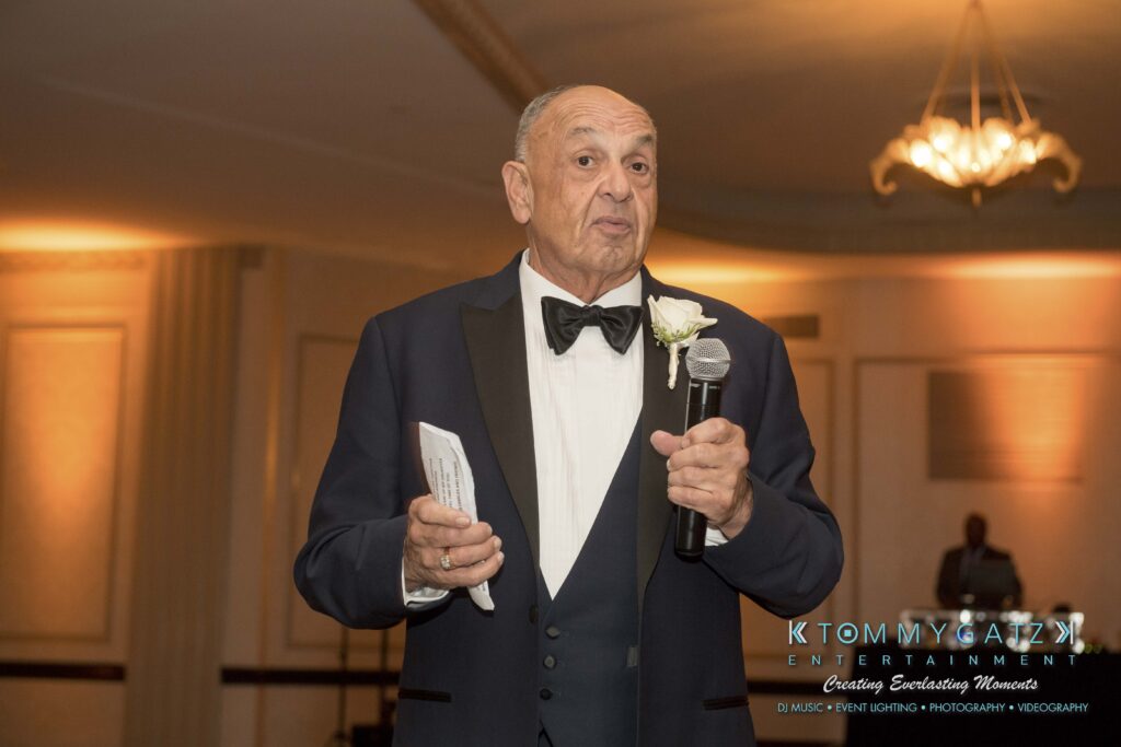 An elderly man in a tuxedo is holding a microphone and speaking at an indoor event with chandelier lighting.