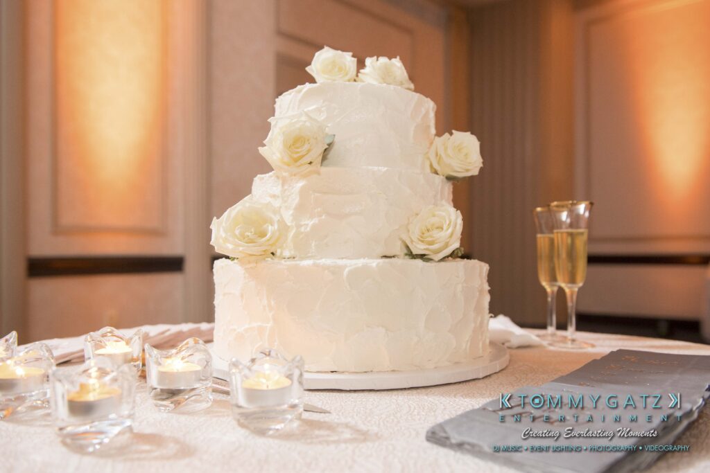 Three-tiered white wedding cake with white roses, surrounded by small candleholders and two champagne glasses on a table.