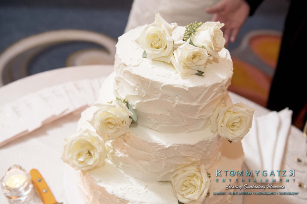 A two-tiered white cake decorated with white roses is placed on a table, with a knife and fork nearby.