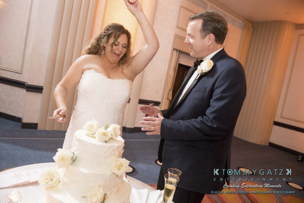 Bride and groom standing near a wedding cake. The bride raises her arm joyfully while the groom looks on. The decorated cake is on a table with an empty champagne flute nearby.