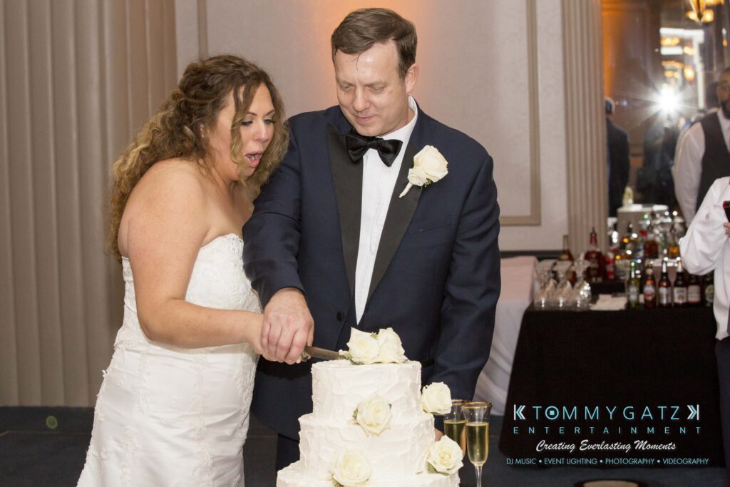 A bride and groom cut a white wedding cake adorned with roses at a reception.