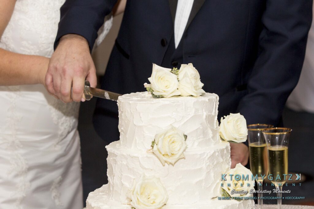 A couple in formal attire cuts a three-tier white cake decorated with white roses, next to two filled champagne glasses.