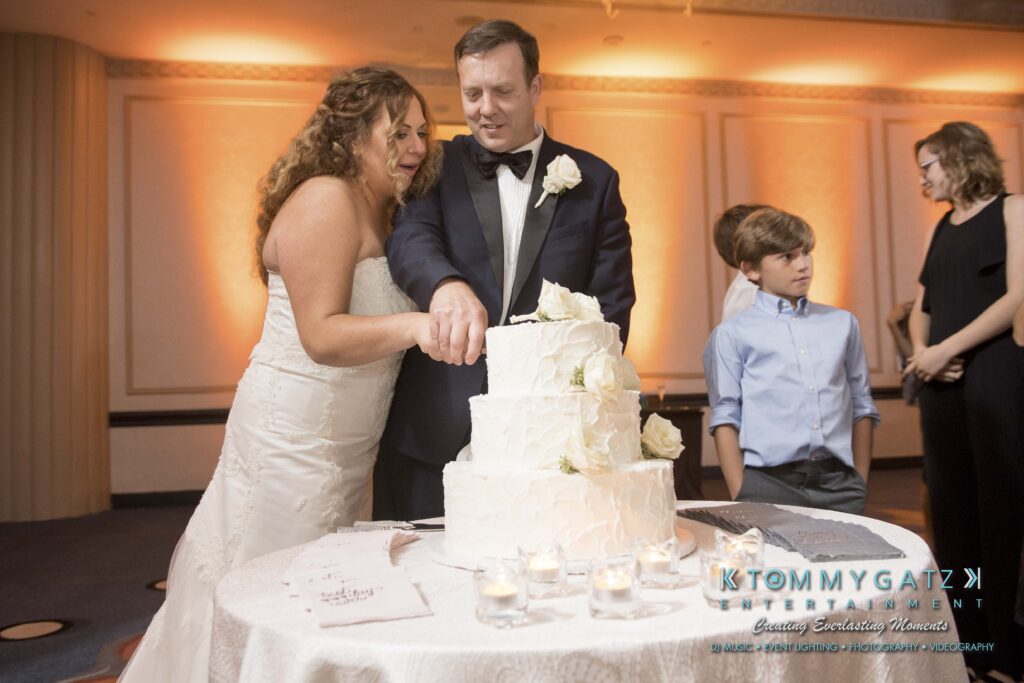 A bride and groom in formal attire cut a three-tiered white wedding cake together. A boy stands nearby, and a woman is in the background. Candles are on the table.