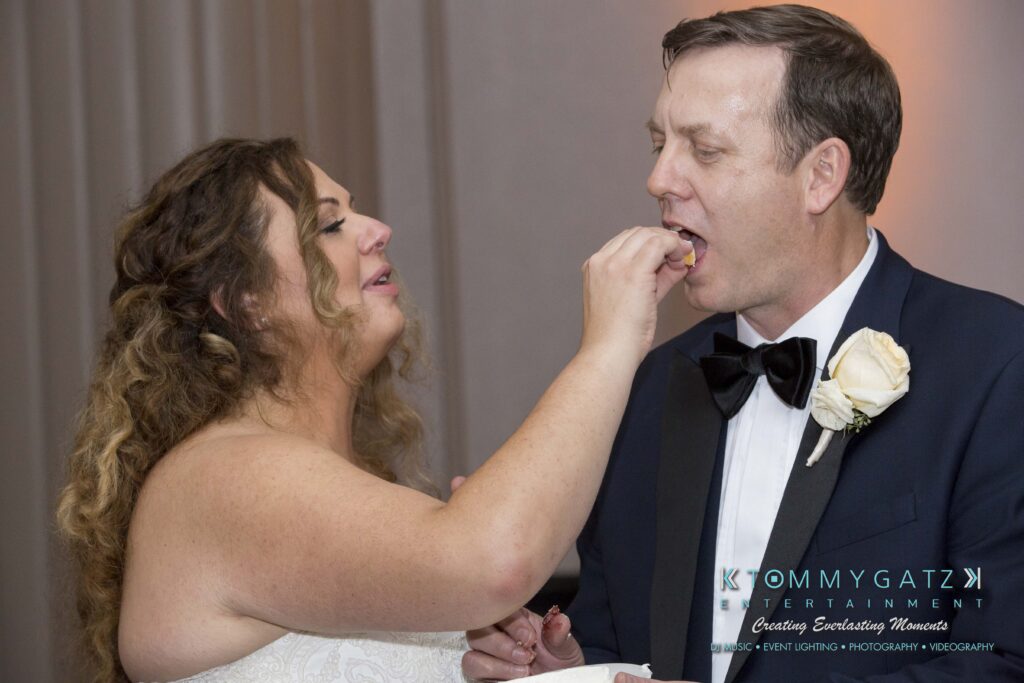 A bride feeds cake to a groom at a wedding reception, both smiling. They are dressed in formal attire.