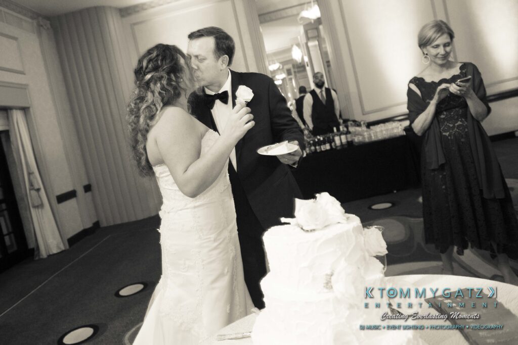 Bride and groom kissing at their wedding reception, holding cake and plates. A guest stands nearby using her phone.