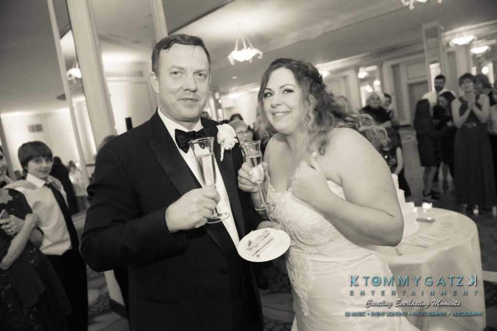 A bride and groom in formal attire hold champagne glasses and a plate, smiling at their wedding reception indoors.