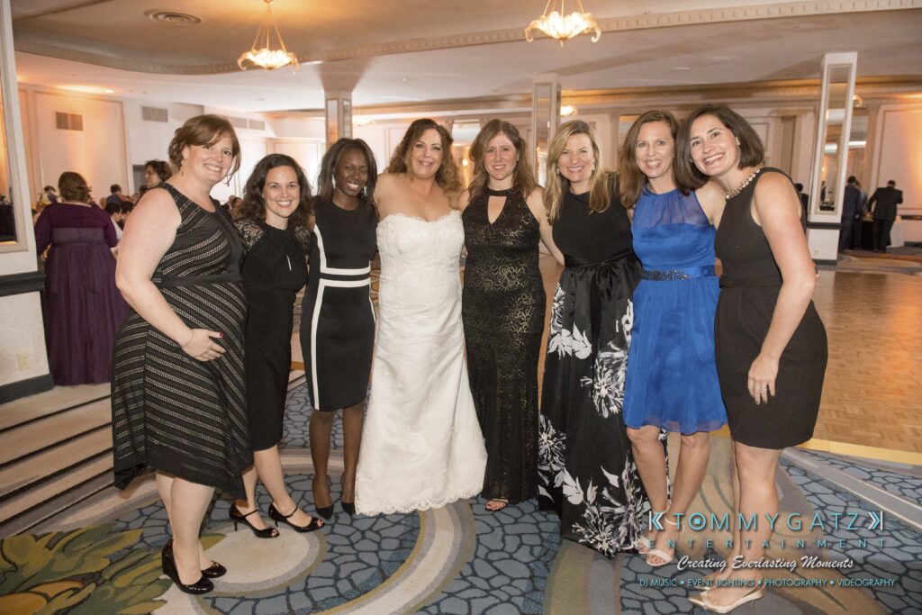 A bride in a white gown stands with seven women wearing various dresses in a formal event setting with ornate carpeting and chandeliers.