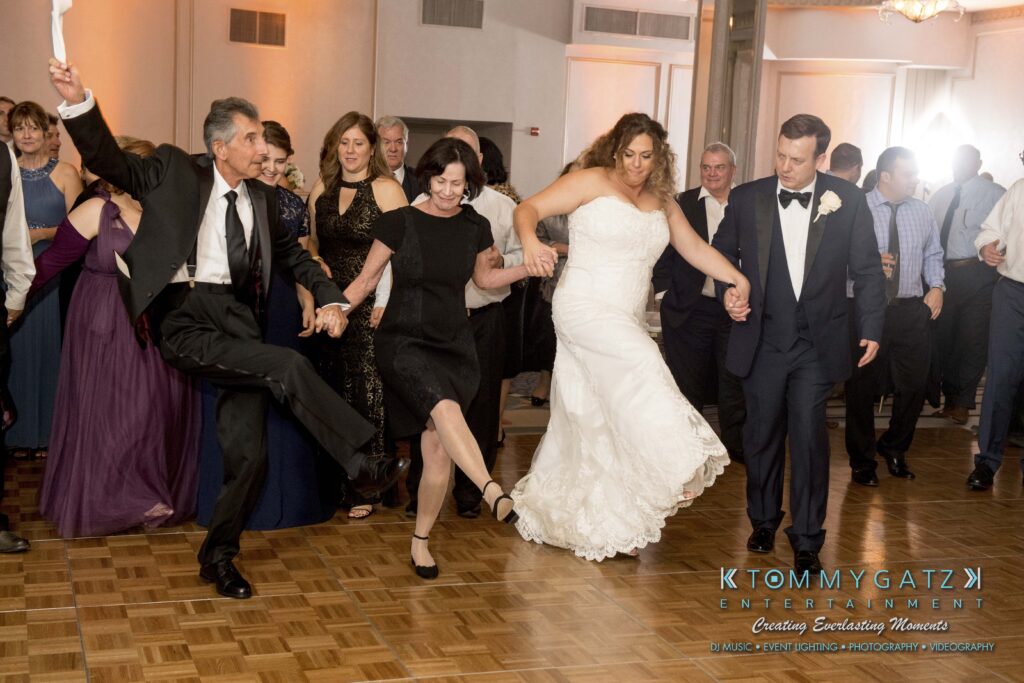 A group of people, including a bride and groom, are dancing in a circle at a wedding reception. They hold hands and kick one leg in the air, smiling and enjoying the celebration.