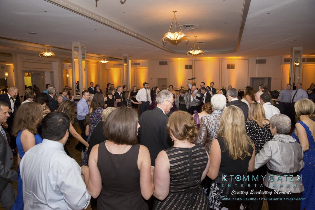 A large group of people stand and talk at a formal event in a spacious ballroom with chandeliers and ambient lighting.