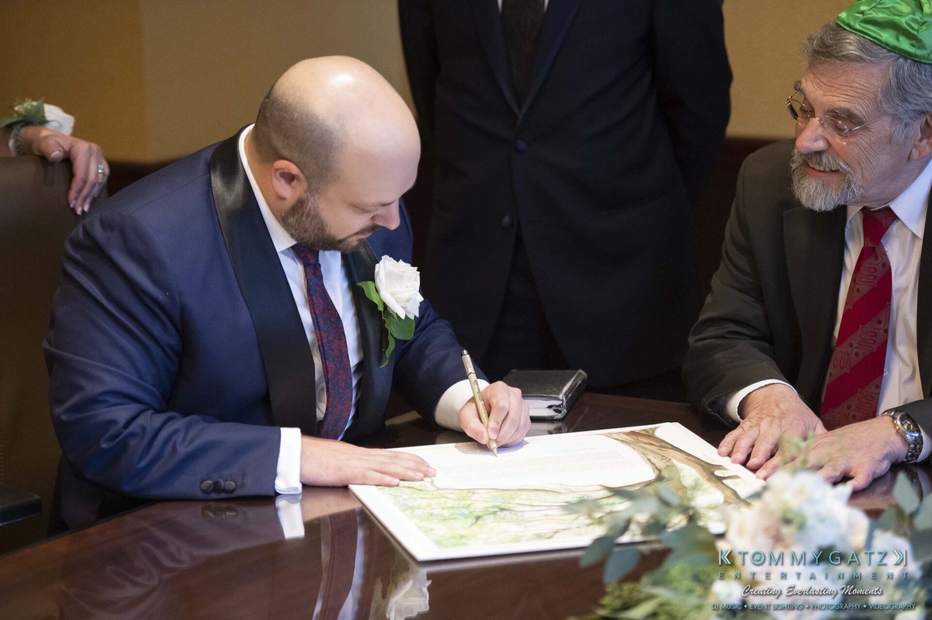 A person in formal attire is signing a document at a table, surrounded by others in similar attire.
