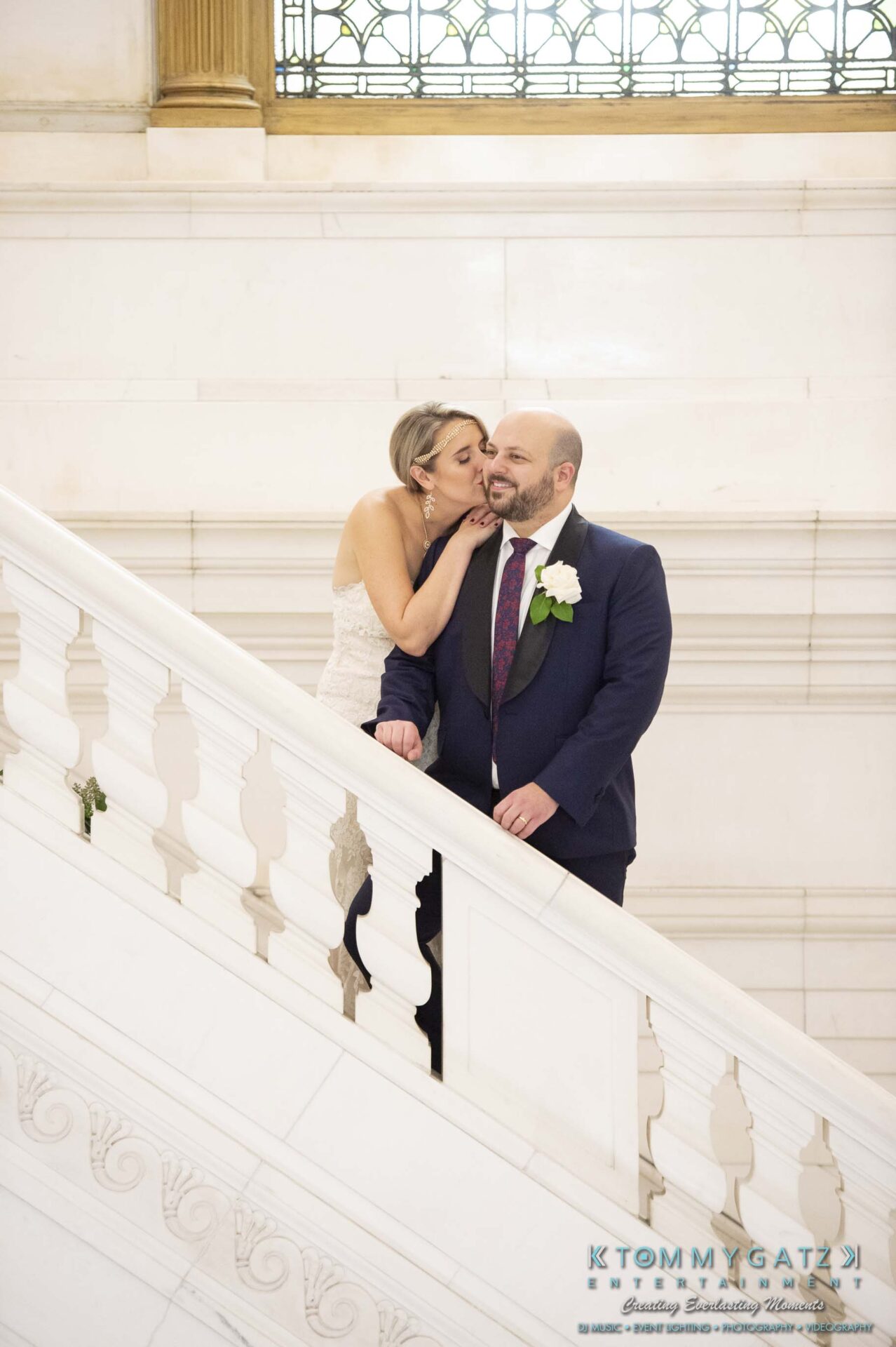 A bride kisses the cheek of a smiling groom on a marble staircase.