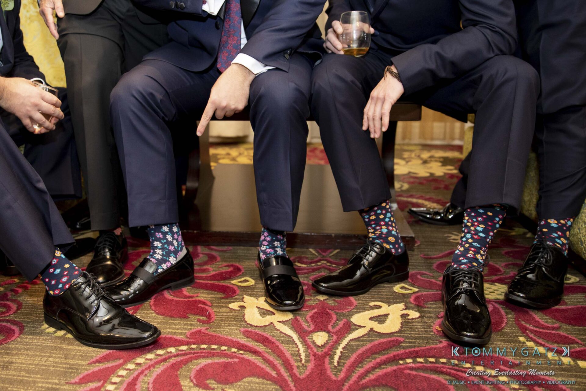 Group of three men seated, showing off colorful patterned socks and shiny black dress shoes on a patterned carpet.