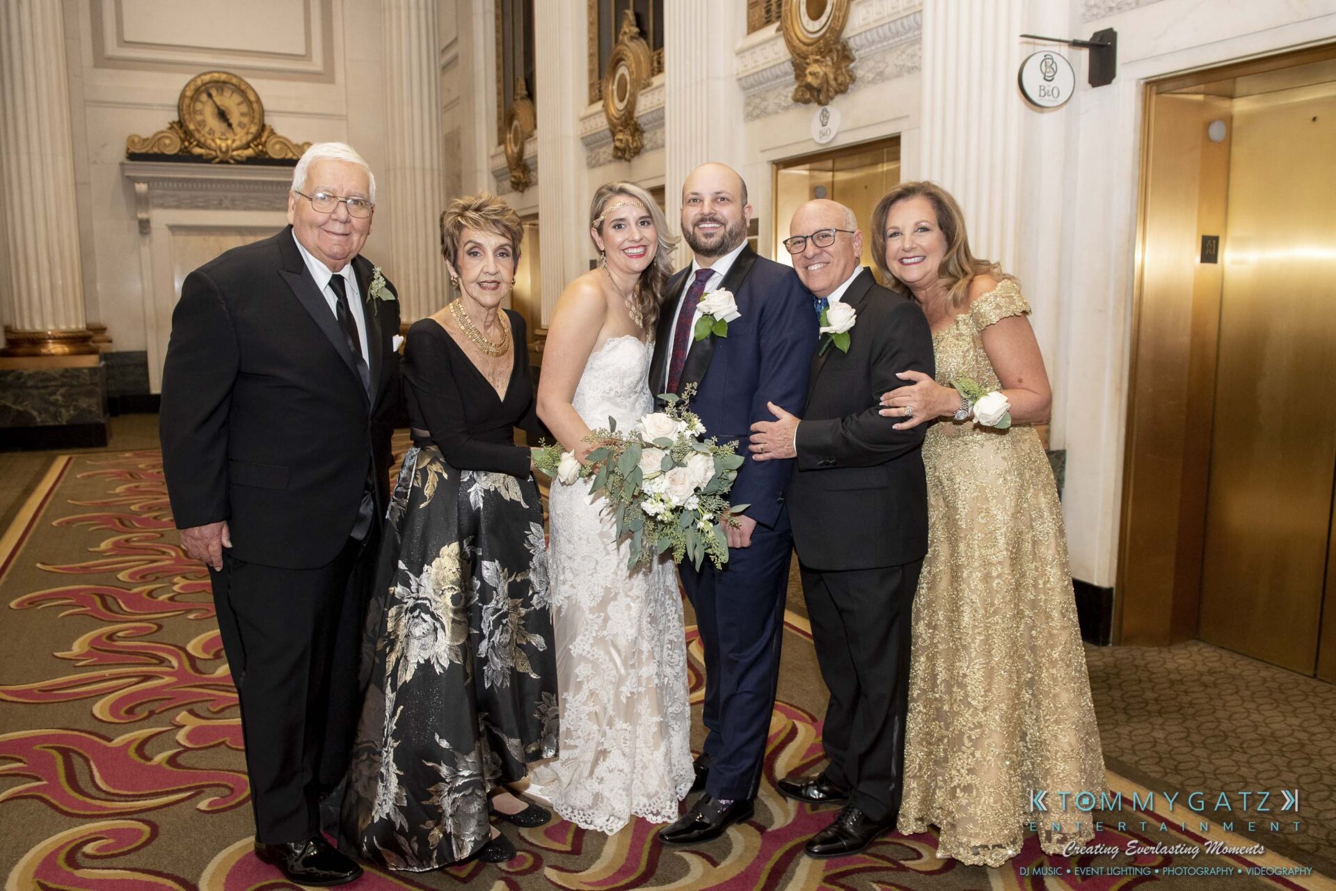 A bride and groom stand with four older adults in formal attire inside a grand building with ornate decor.