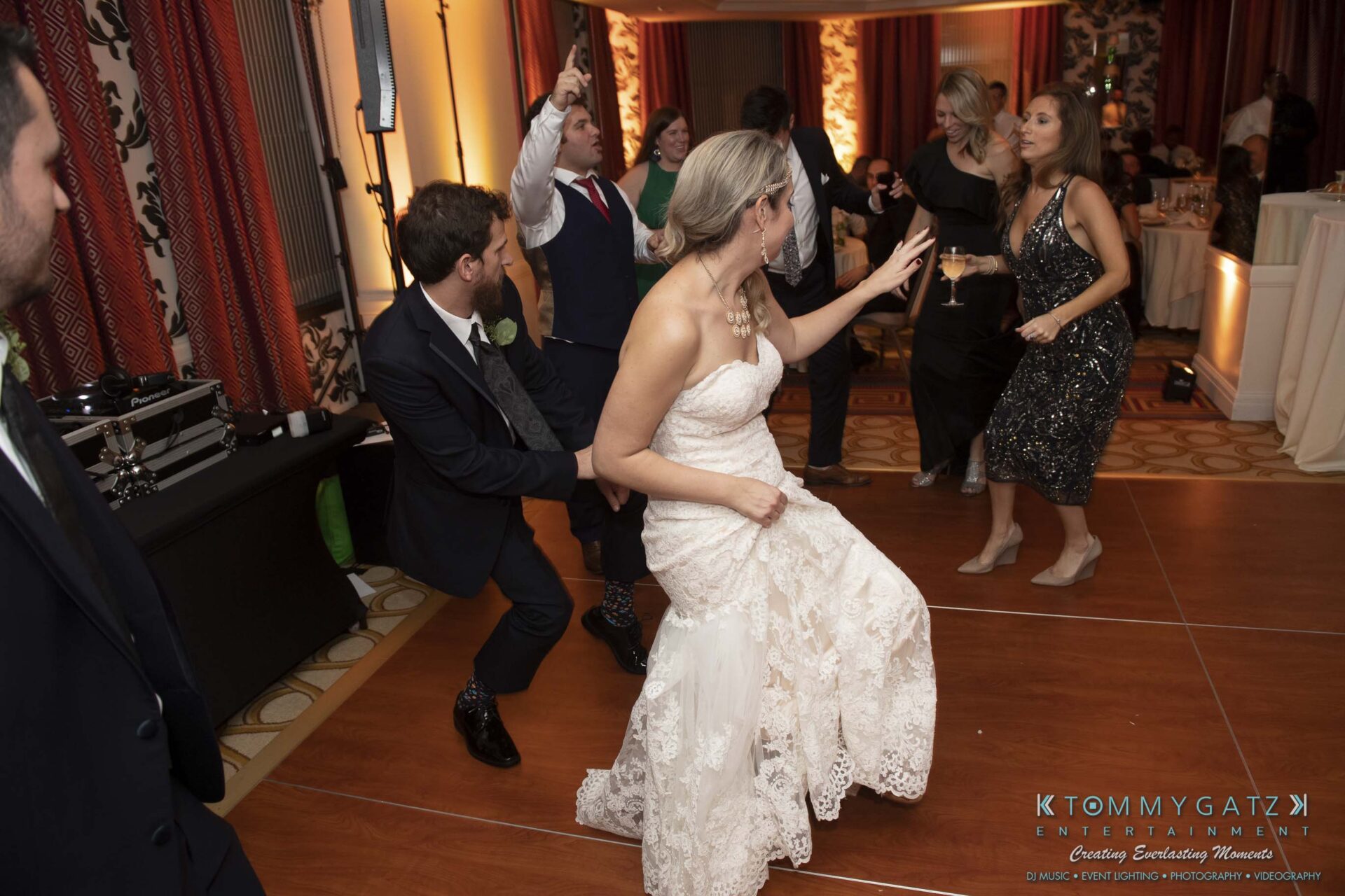 People in formal wear are dancing at a lively indoor event with orange lighting. A bride in a white gown is at the center.