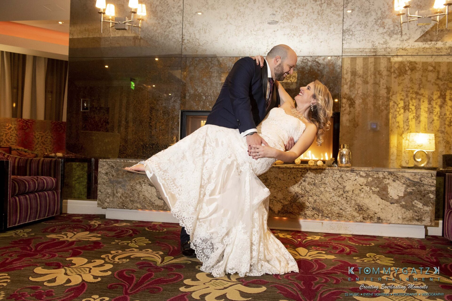 A groom in a suit joyfully lifts his bride in a lace wedding dress in a hotel lobby with ornate decor.