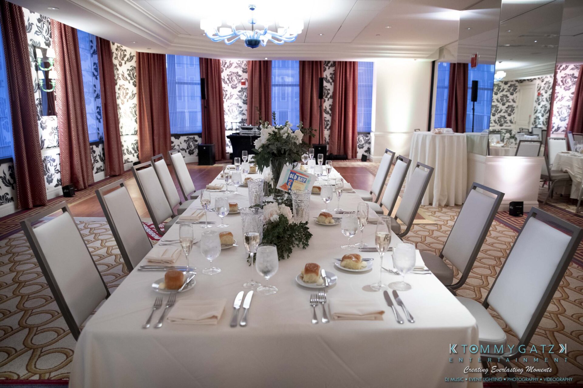 A long dining table set for a formal event in an elegant room, with white tablecloth, arranged cutlery, glasses, and rolls on plates. The room has patterned walls, curtains, and a large chandelier.