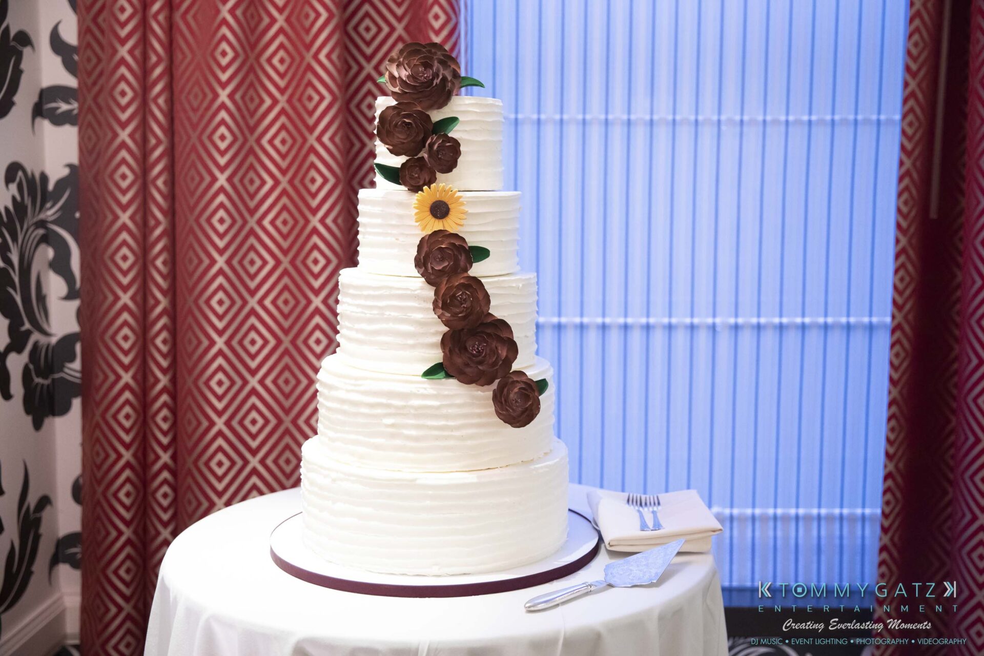 Four-tier white wedding cake with brown flower decorations and a single yellow flower, placed on a table with a fork and knife nearby.