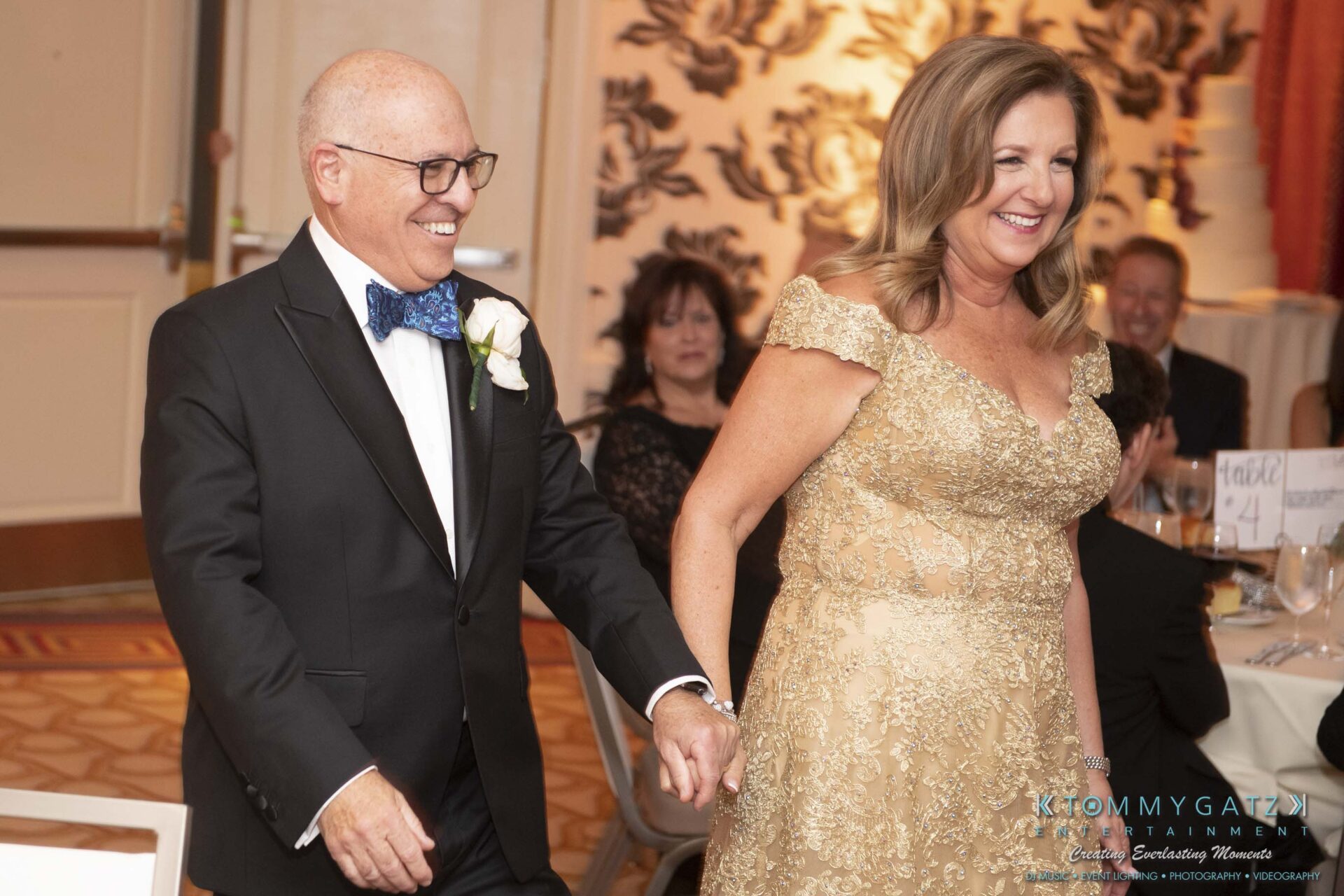A couple in formal attire is smiling and holding hands while walking, with seated guests visible in the background at an event.
