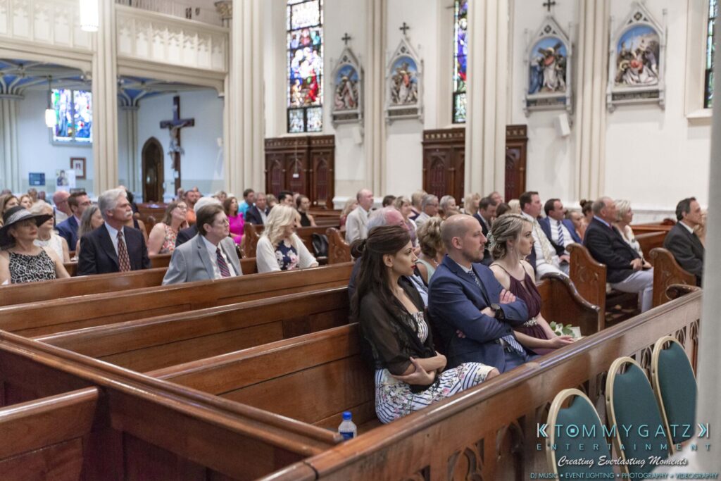 People seated in wooden pews inside a church with stained glass windows and decorative arches.