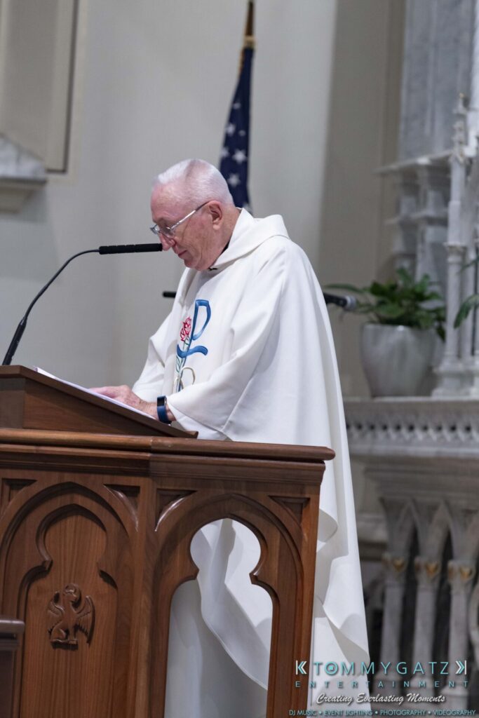 A man in a white robe is speaking at a podium in a church, with an American flag and plant visible in the background.