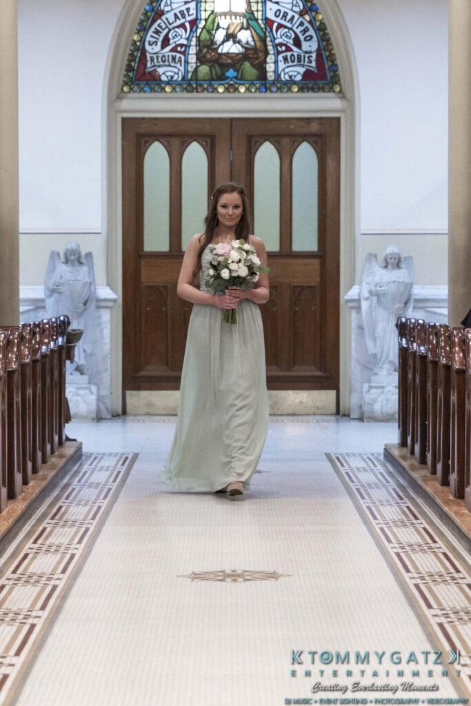 A woman in a light green dress holds a bouquet while walking down a church aisle lined with wooden pews.