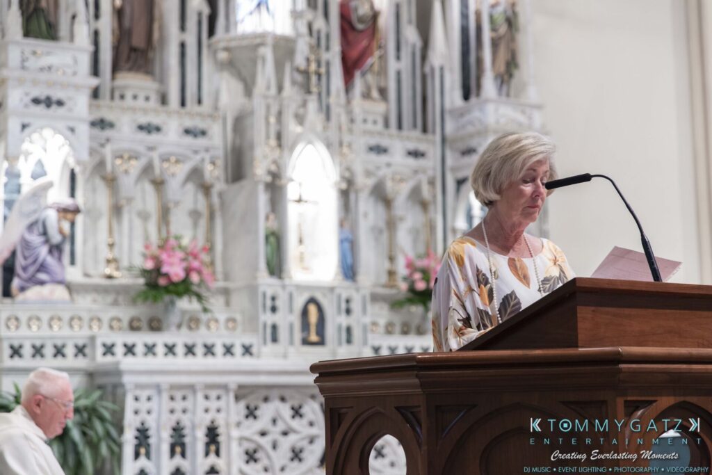 A woman reads at a lectern in a church, with an elderly man seated nearby. The altar is ornately decorated.