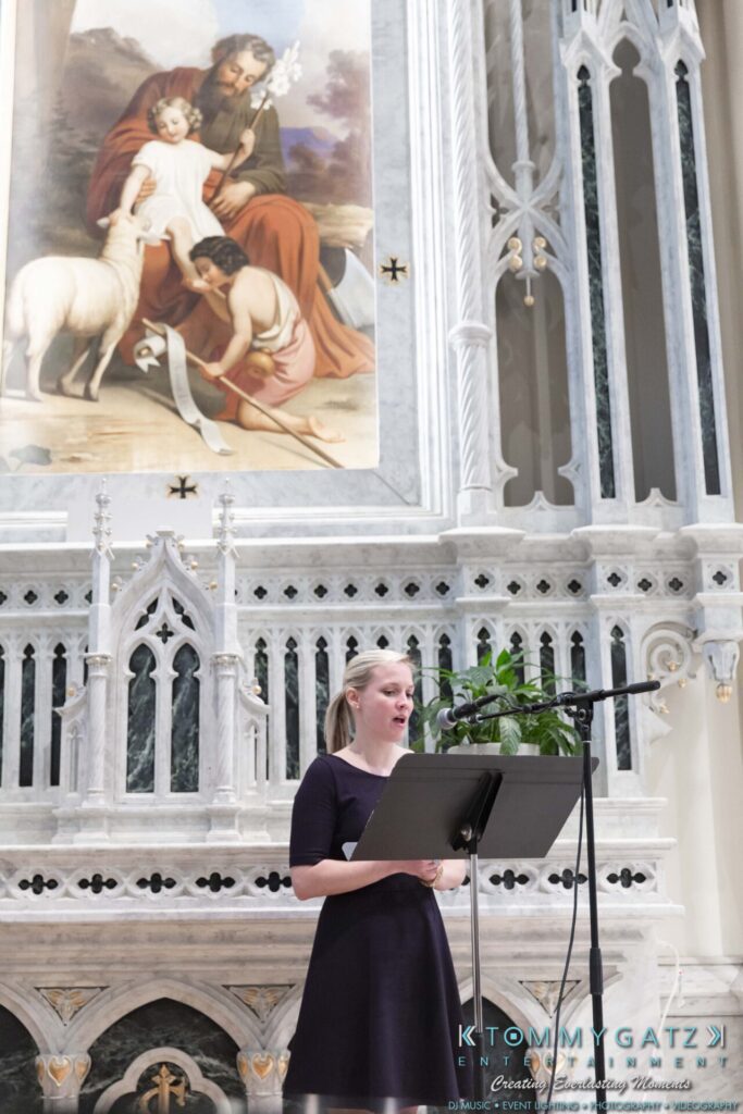 A woman stands at a podium speaking, with a microphone in front of her. Behind her is an ornate altar and a large religious painting depicting Jesus, a child, and a lamb.