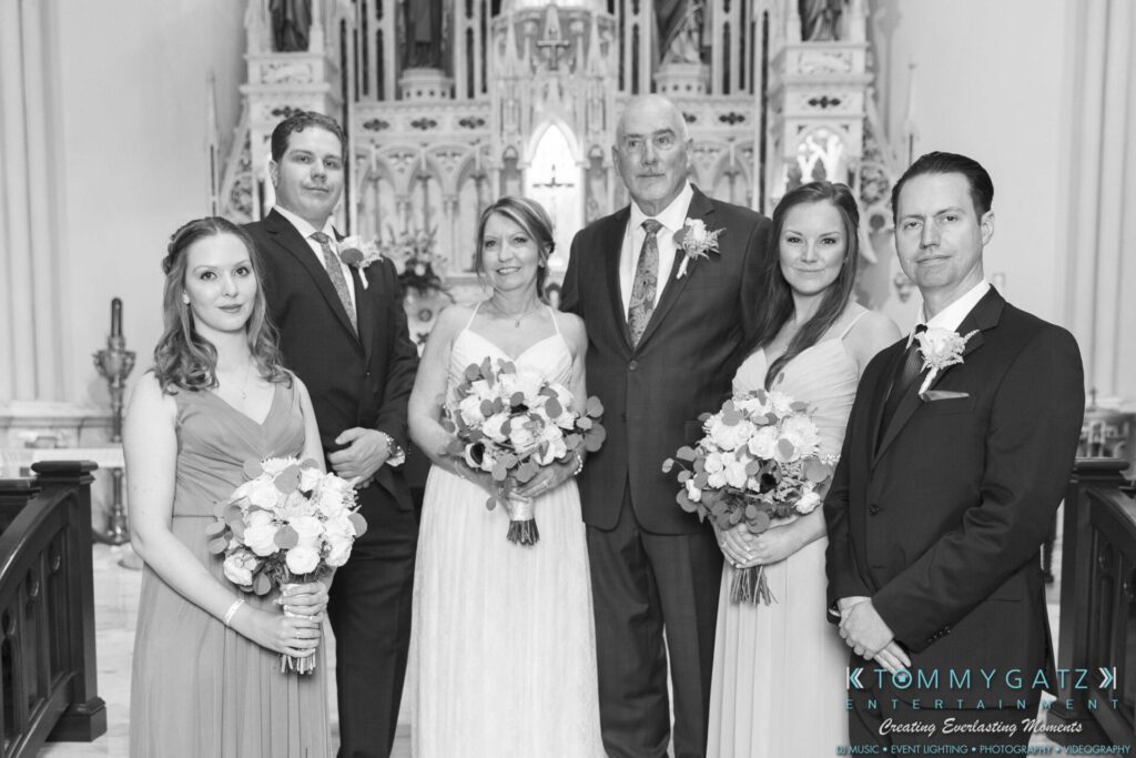 A wedding party poses in a church, featuring a bride, groom, two bridesmaids, and two groomsmen holding bouquets.