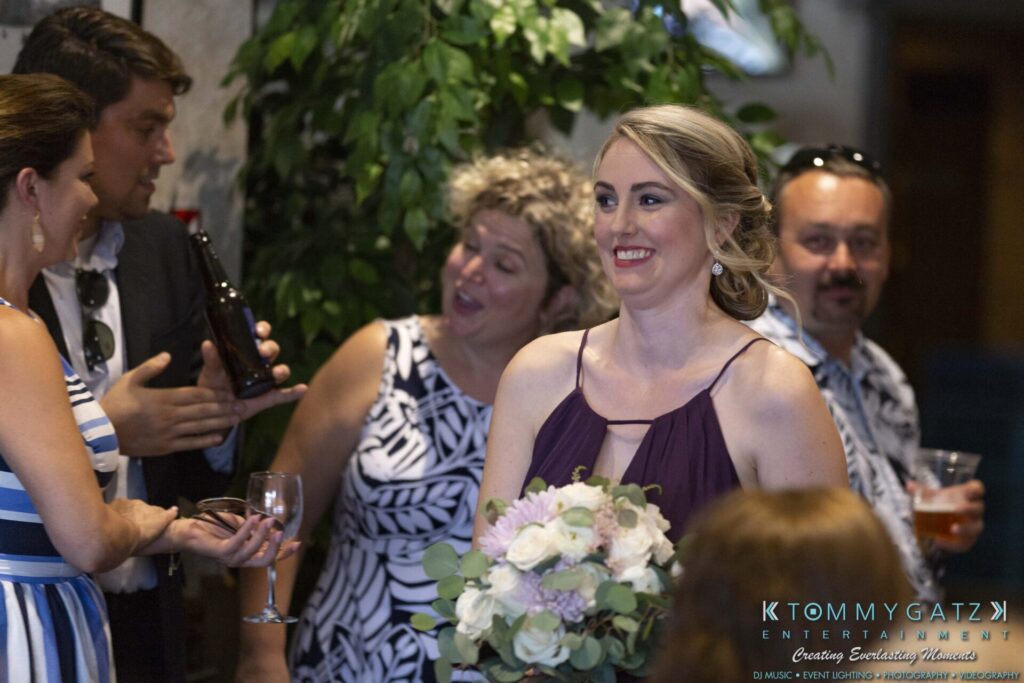 A woman in a purple dress holds a bouquet of flowers at a social gathering. People around her are engaged in conversation and holding drinks.