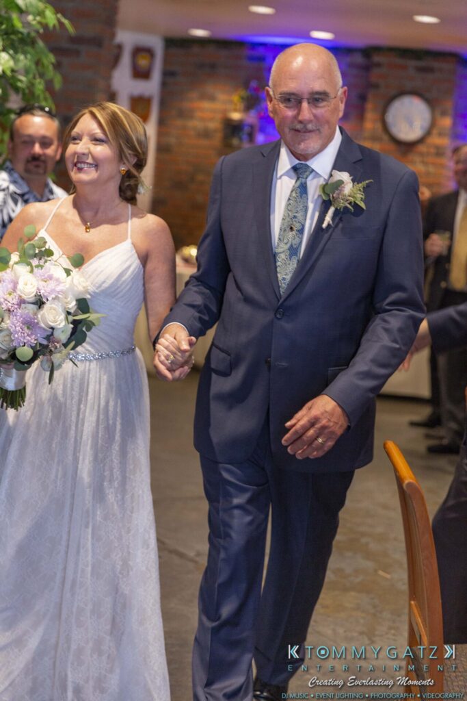 A couple in formal attire walks hand in hand indoors. The woman holds a bouquet, and both appear to be smiling.