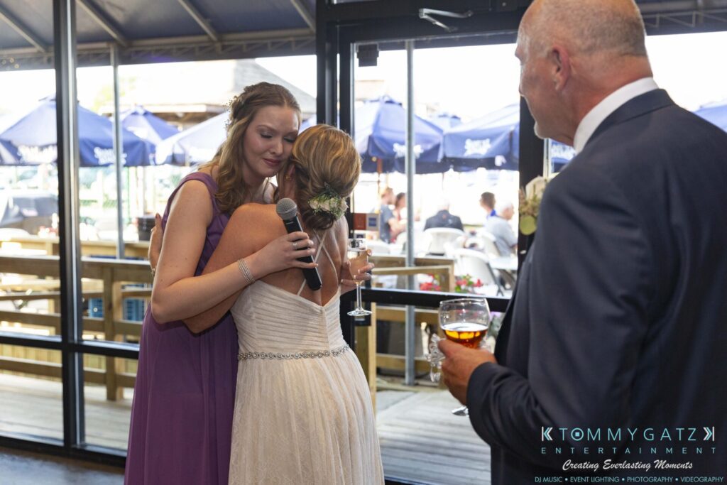 A bridesmaid in a purple dress gives a speech and embraces the bride, who holds a drink. A man in a suit observes them, holding a glass, with people visible outside on a patio.