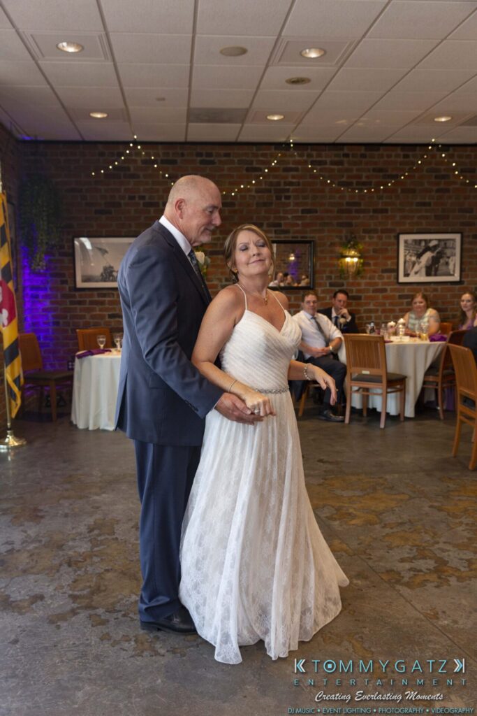 A couple, dressed in formal attire, dances at an indoor reception with guests seated at tables in the background.