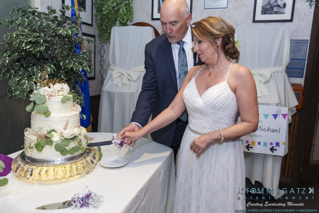 A couple in formal attire cuts a three-tiered white cake decorated with greenery and flowers at an indoor event.