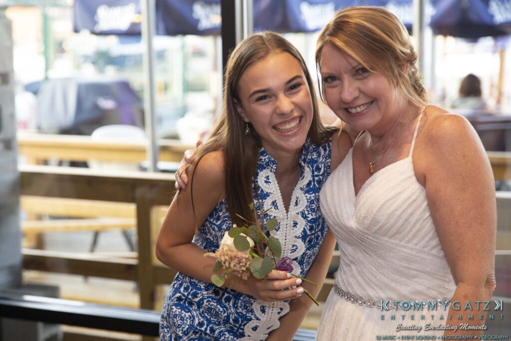 A young woman in a blue dress and an older woman in a white dress smile while posing together indoors. The younger woman holds a small bouquet.