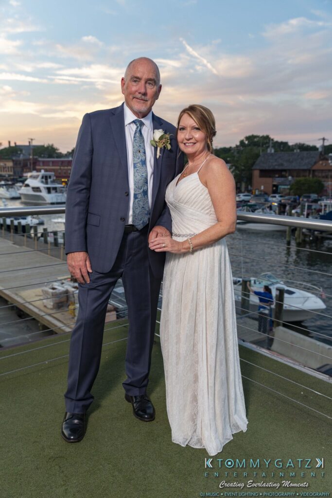 A couple stands on a deck overlooking a marina. The man wears a suit and the woman is in a white dress.