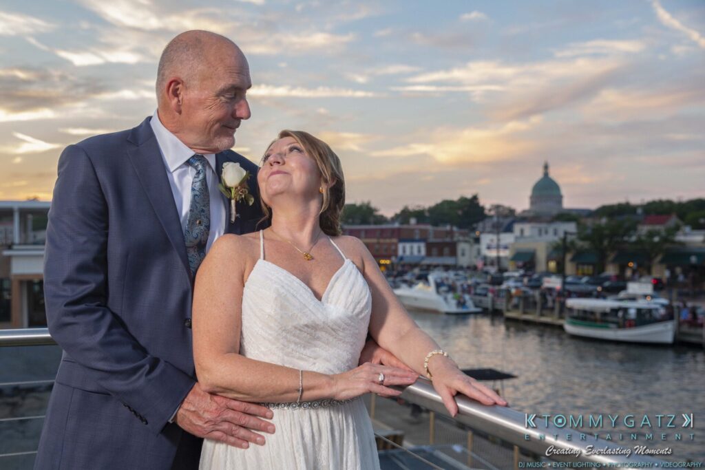 A couple in wedding attire stand on a balcony overlooking a waterfront at sunset.