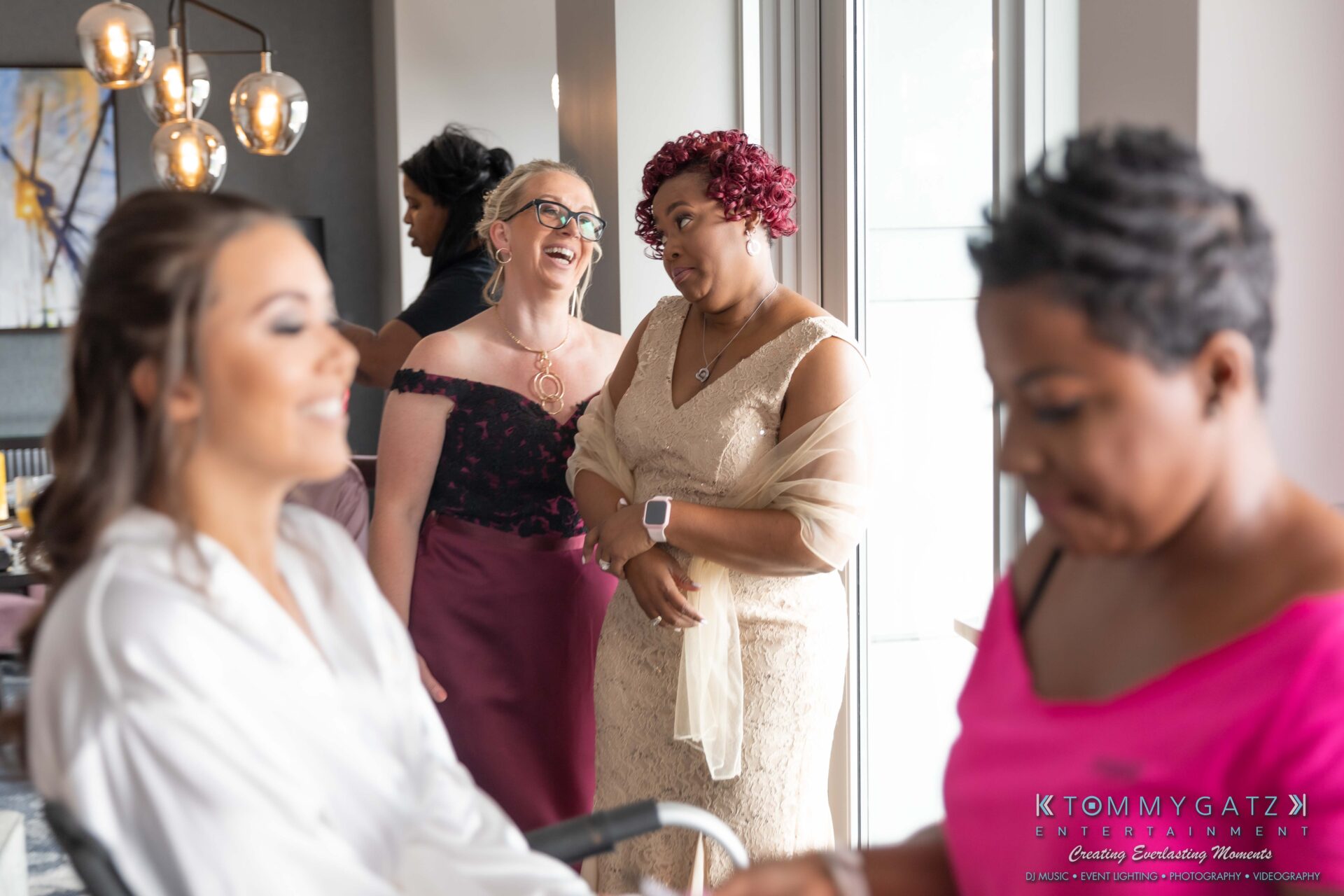 Two women in formal dresses watch another woman getting ready, while a fourth woman assists. The setting appears to be a preparation area, possibly for an event.