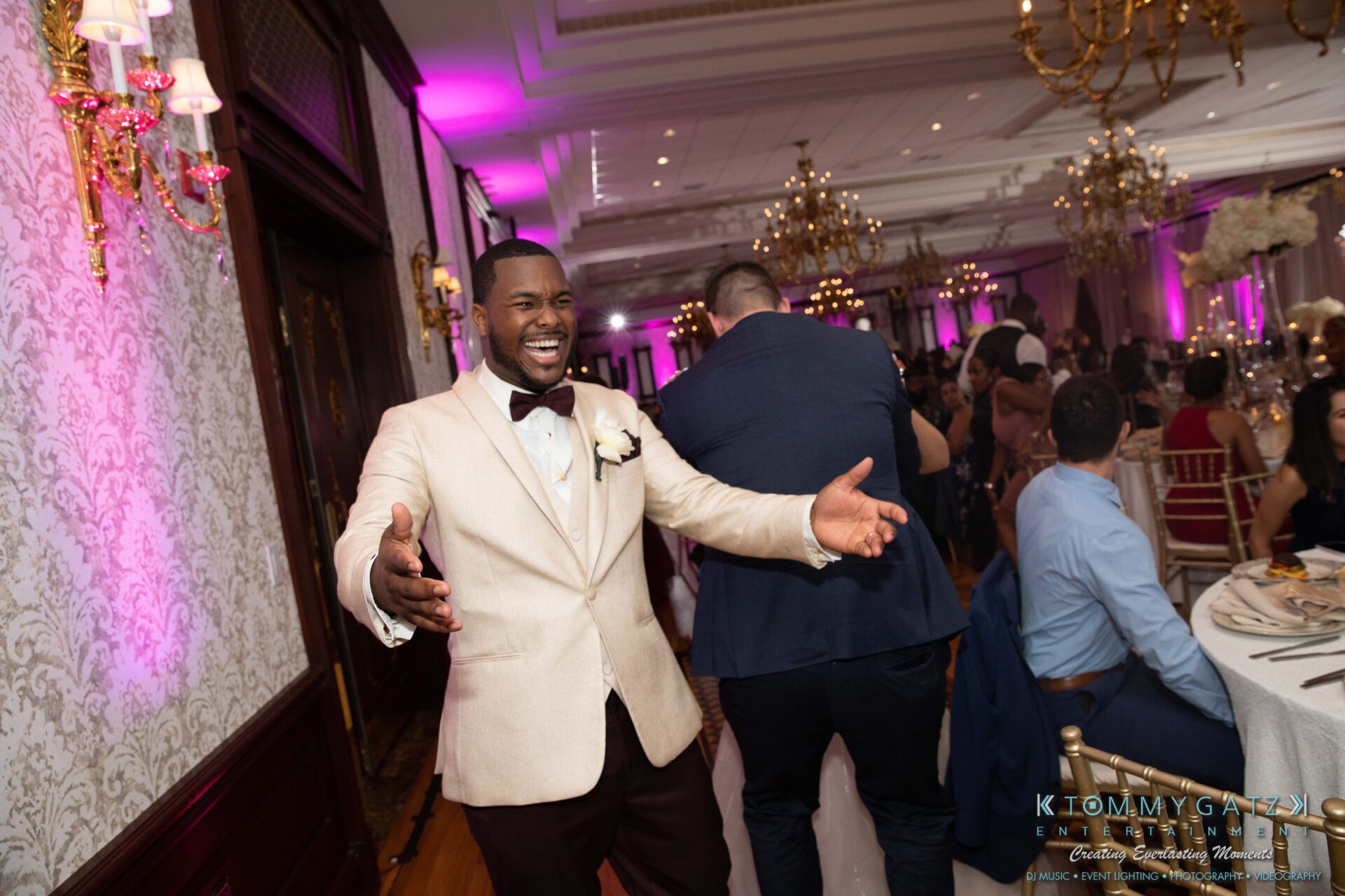 A smiling man in a beige suit with a bow tie stands with open arms at an indoor event with chandeliers and decorated tables.