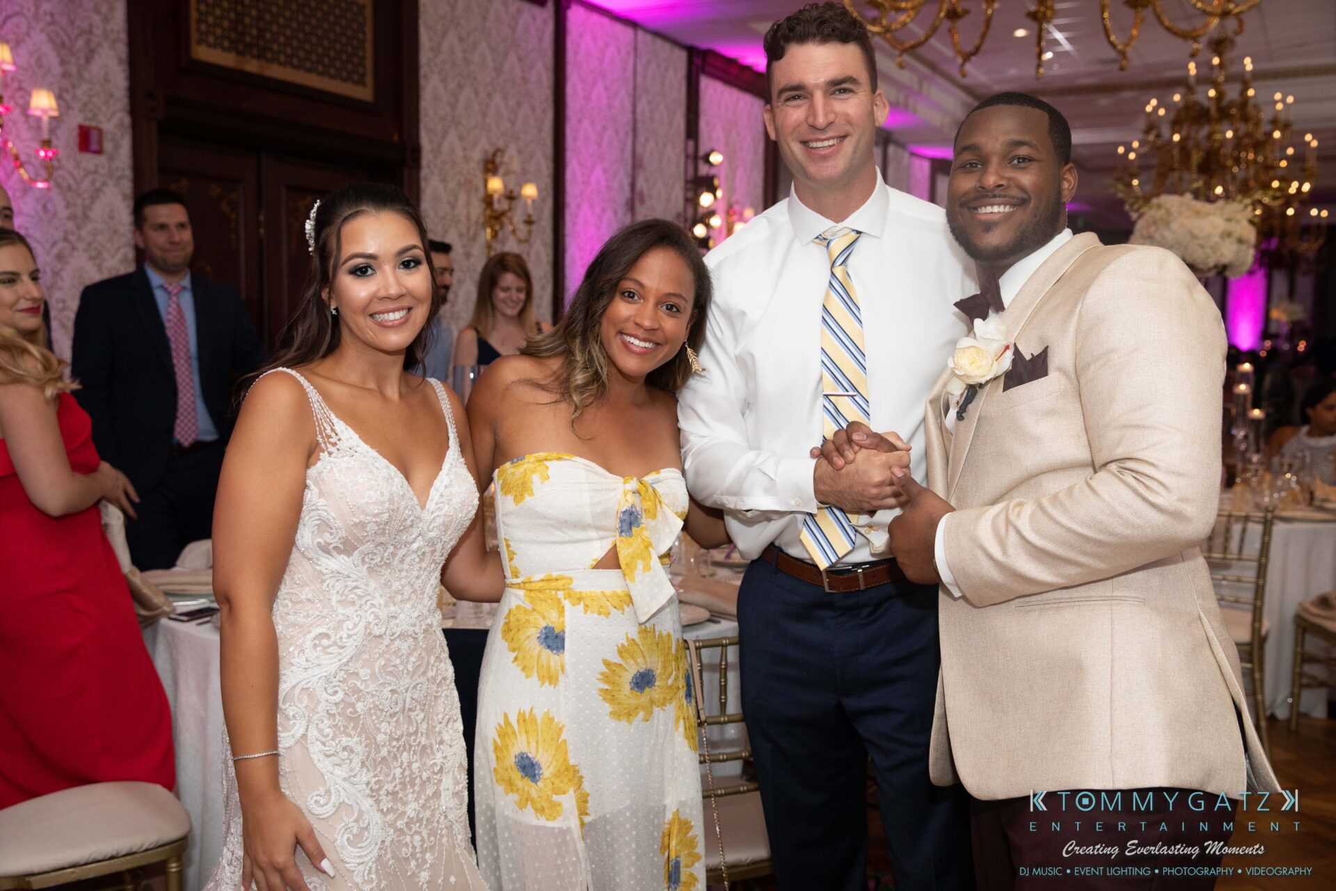 A group of four people, three women and a man, smiling and standing together at a formal event. The room is decorated, and guests are visible in the background.